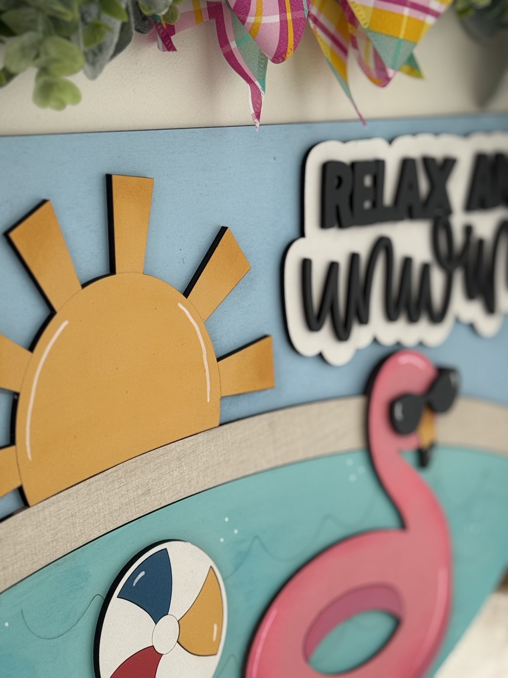 A decorative summer sign featuring a yellow sun, a pink flamingo float in water, a colorful beach ball, and the phrase RELAX AND UNWIND in bold letters with festive ribbon at the top.