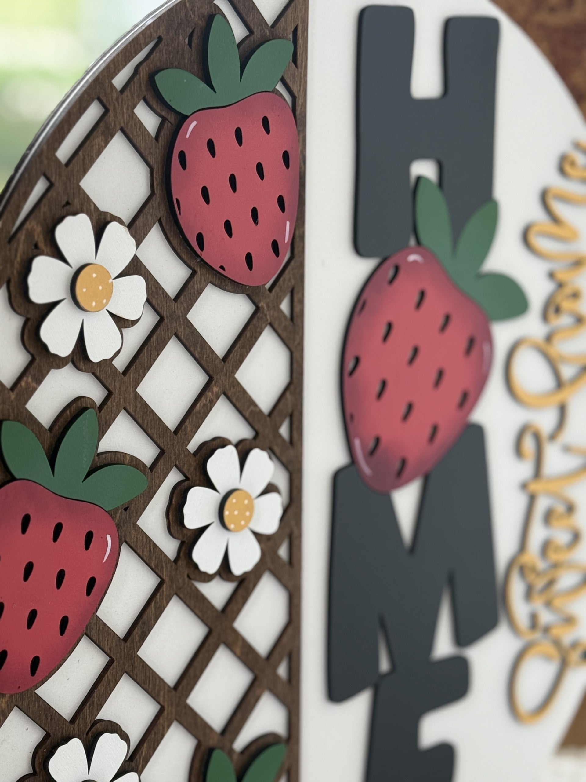 A decorative sign featuring strawberries and white flowers on a wooden lattice next to large black letters spelling HOME, with part of the word always visible in yellow script.
