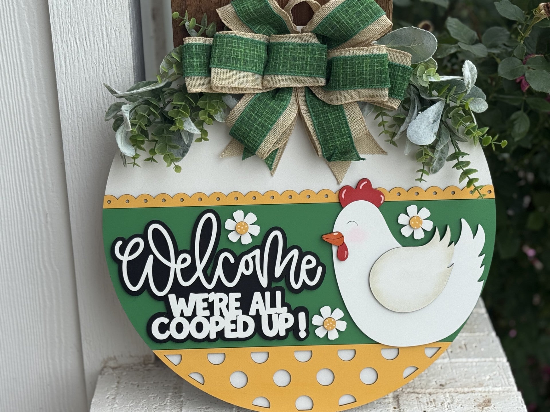 A round decorative sign with a cartoon chicken, flowers, and the text Welcome Were All Cooped Up! It features a green and yellow polka-dot pattern and is topped with a green and burlap bow and artificial greenery.