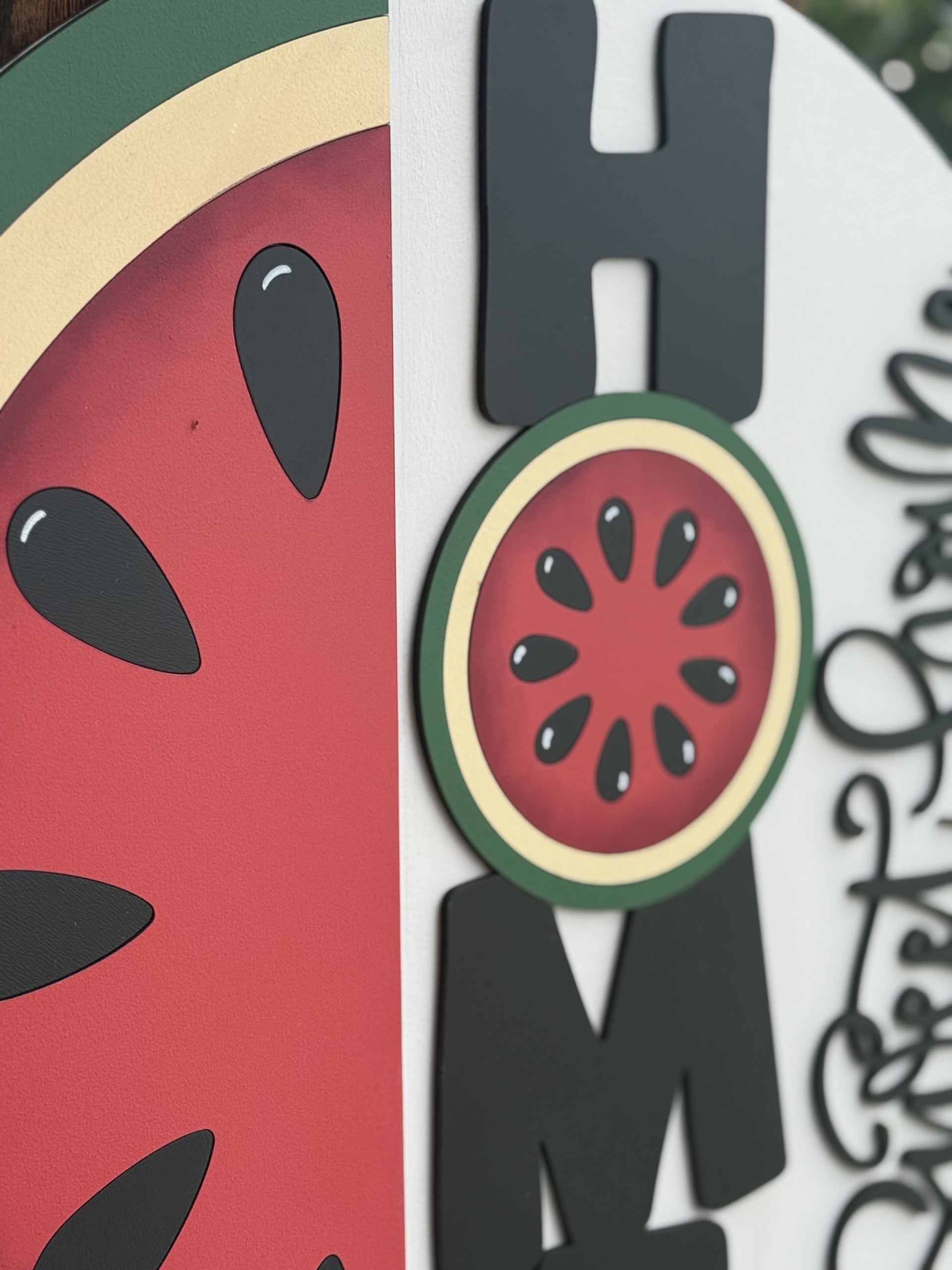 A close-up of a decorative sign featuring the word HOME with a watermelon slice used as the letter O, and part of a handwritten Welcome phrase visible on the right side.