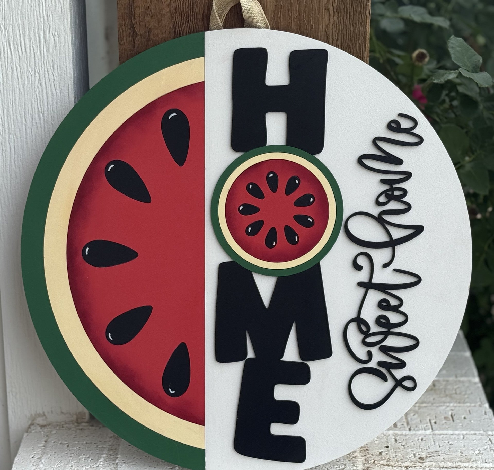 A round sign features a watermelon slice on the left and the word HOME vertically, with the O as a watermelon. Beside it, sweet home is written in cursive on a white background. Greenery is visible in the background.