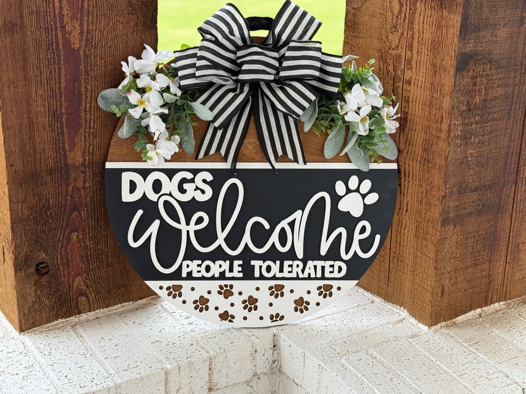 A round sign with a black and white striped bow, flowers, and greenery reads, Dogs welcome, people tolerated, with paw prints, hanging on a wooden wall.