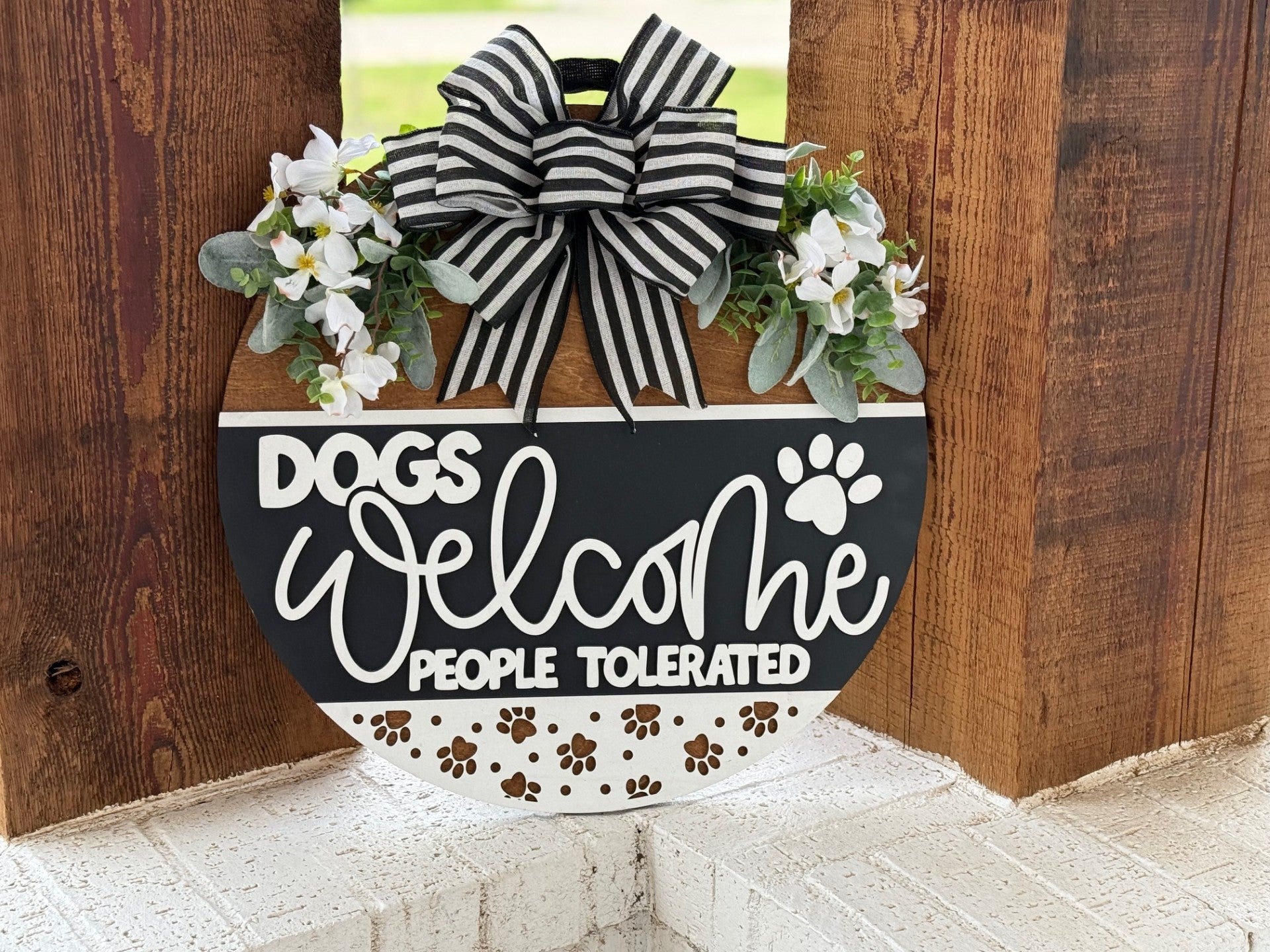 A decorative sign with a striped bow, flowers, and the words Dogs welcome, people tolerated hanging on a wooden wall. The sign also features paw prints along the bottom.