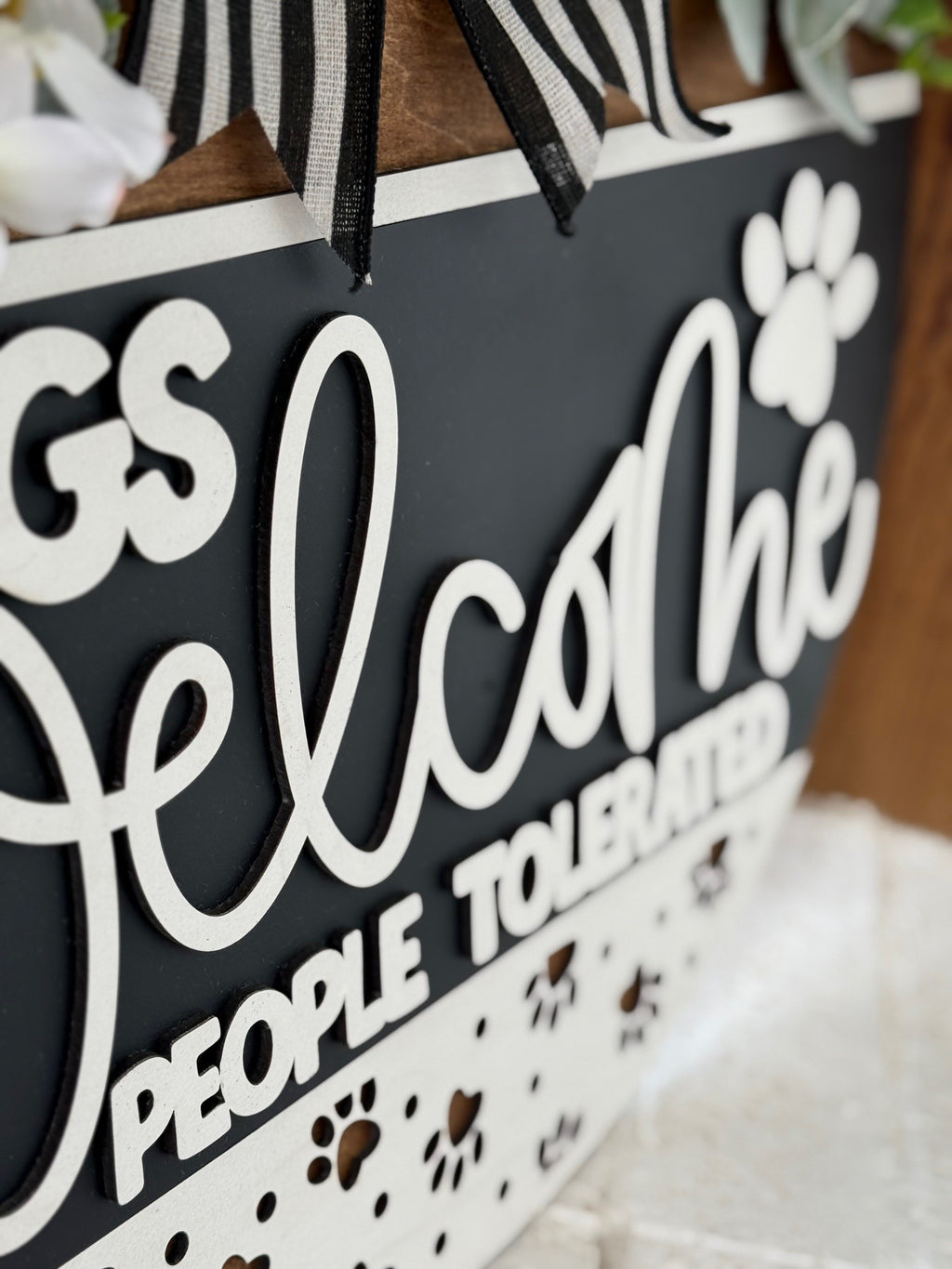 Close-up of a decorative sign with black and white text that reads, Dogs Welcome, People Tolerated, along with paw prints and striped ribbon accents at the top.