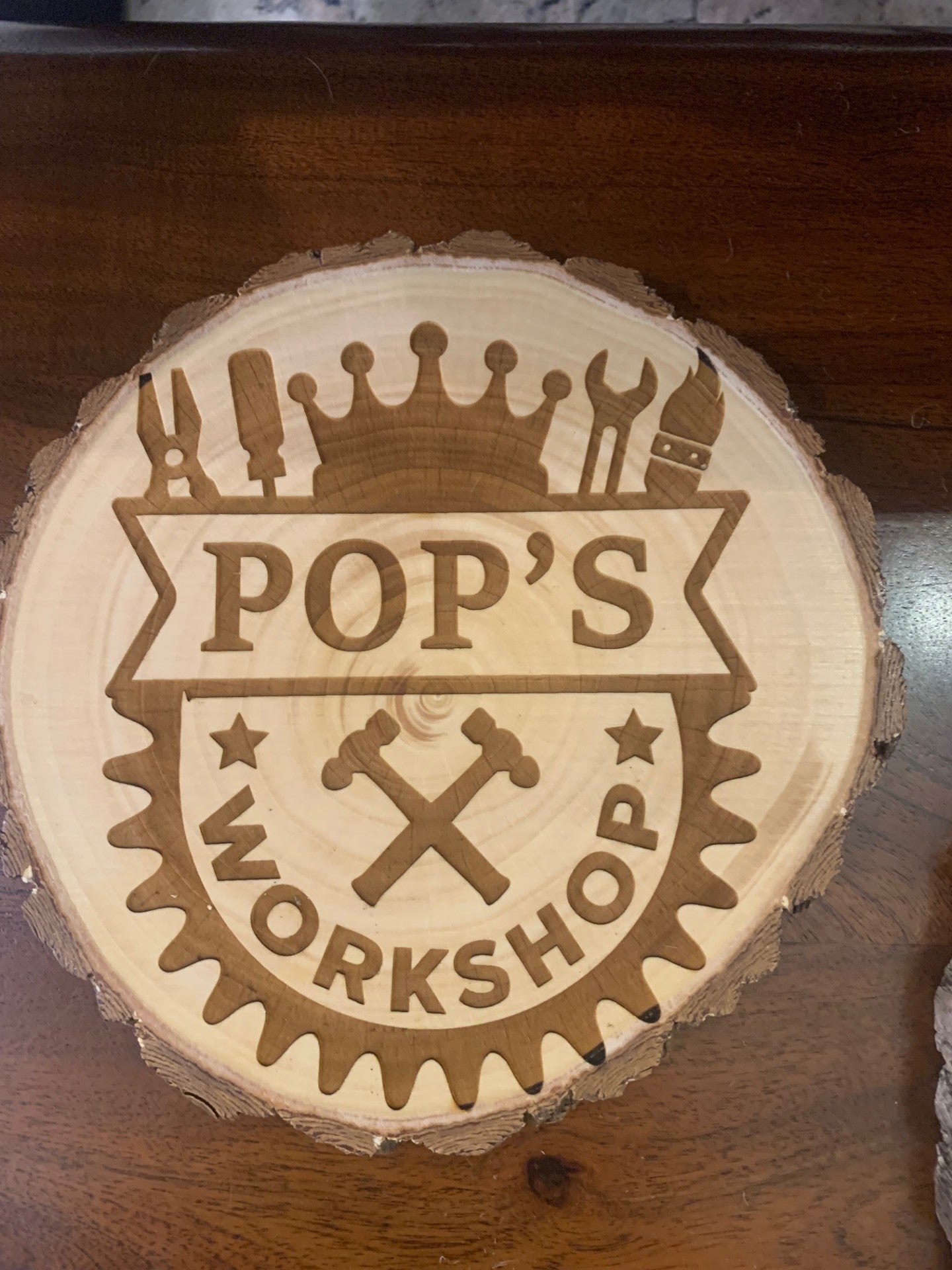 The Dads Workshop or Pops Workshop round wood slice wall decor features engraved drawings of tools, including pliers, screwdriver, wrench, paintbrush, and hammer. DADS WORKSHOP is highlighted with a crown and two crossed hammers beneath it on a rustic wooden plaque.