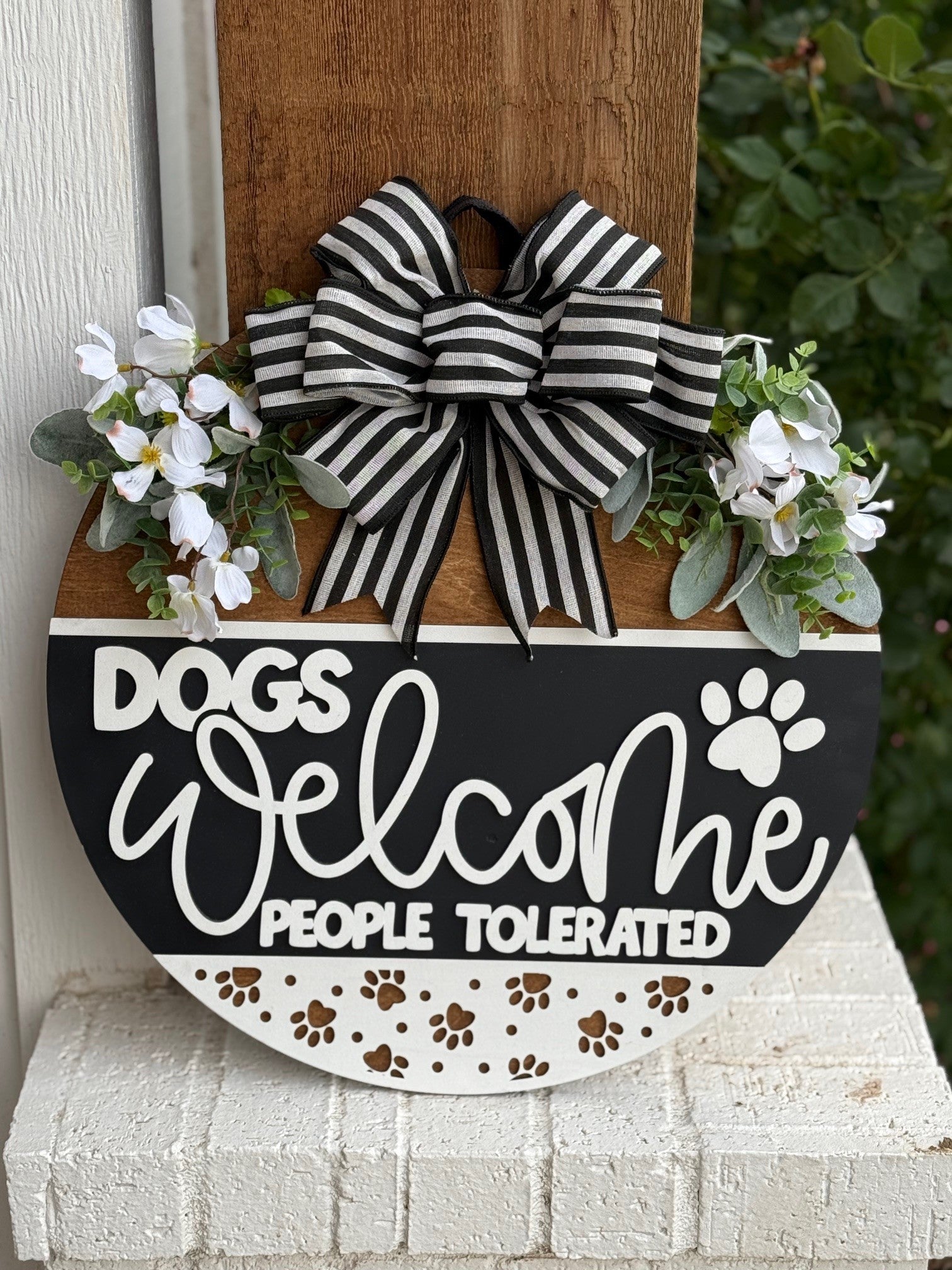 A round sign with white flowers, greenery, and a black-and-white striped bow reads: DOGS WELCOME PEOPLE TOLERATED, with paw prints and a paw graphic on a white, black, and brown background.