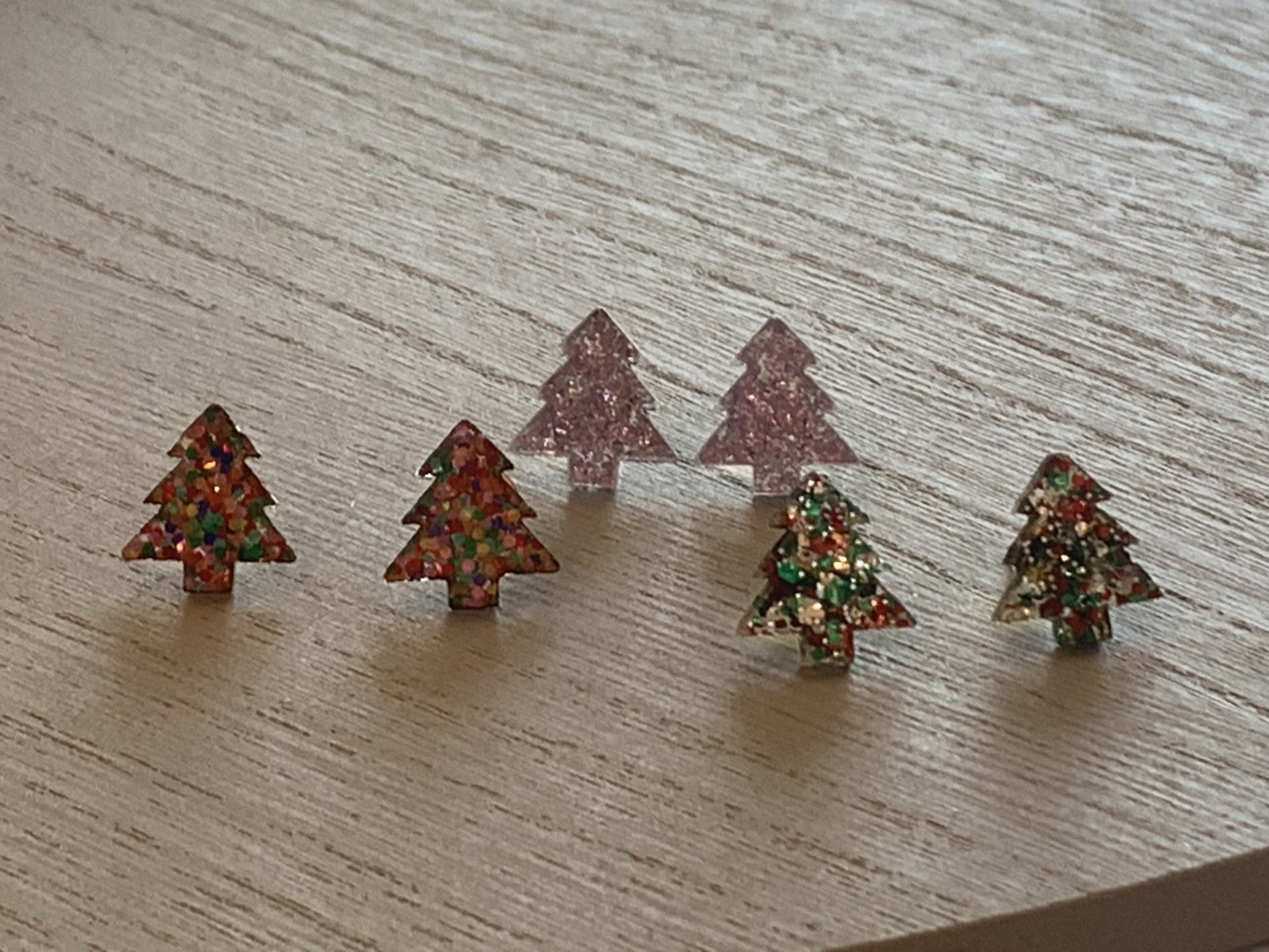 Five Christmas Tree Stud Earrings rest on a light wooden surface: two with confetti textures, one in green and red glitter, and two translucent trees glowing pinkish-purple under soft shadows.