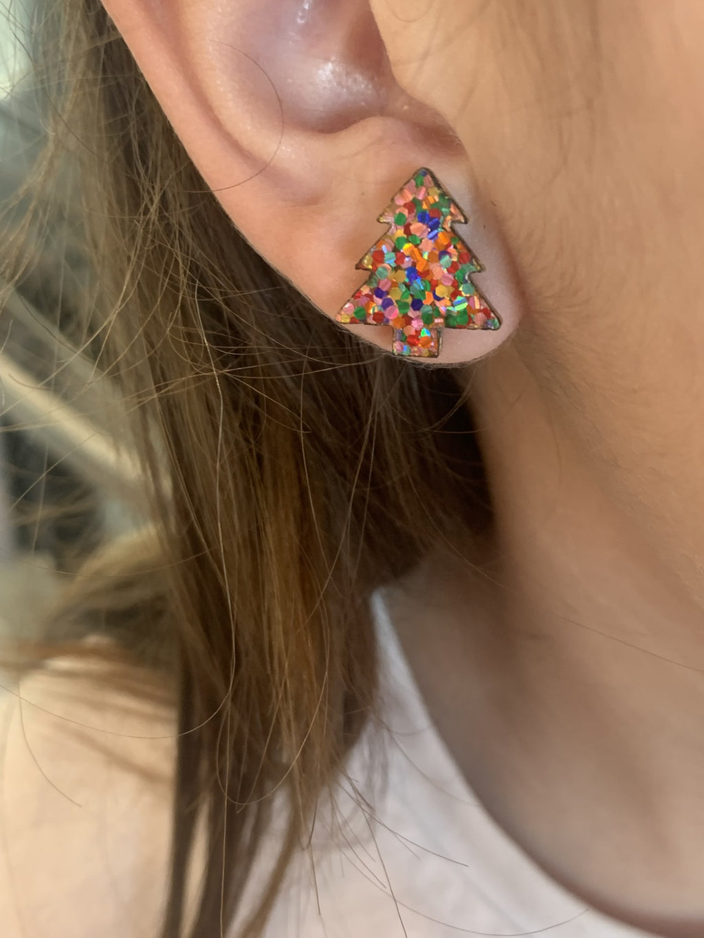 A close-up shows an ear with Christmas Tree Stud Earrings, featuring multicolored specks for a glittery look. Light brown hair partially covers the ear, enhancing the charm.