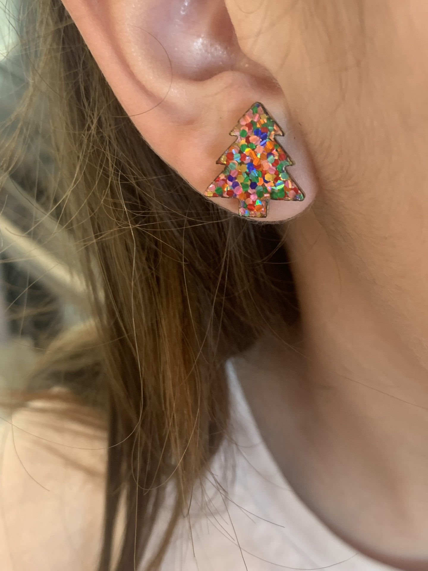 A close-up shows an ear with Christmas Tree Stud Earrings, featuring multicolored specks for a glittery look. Light brown hair partially covers the ear, enhancing the charm.