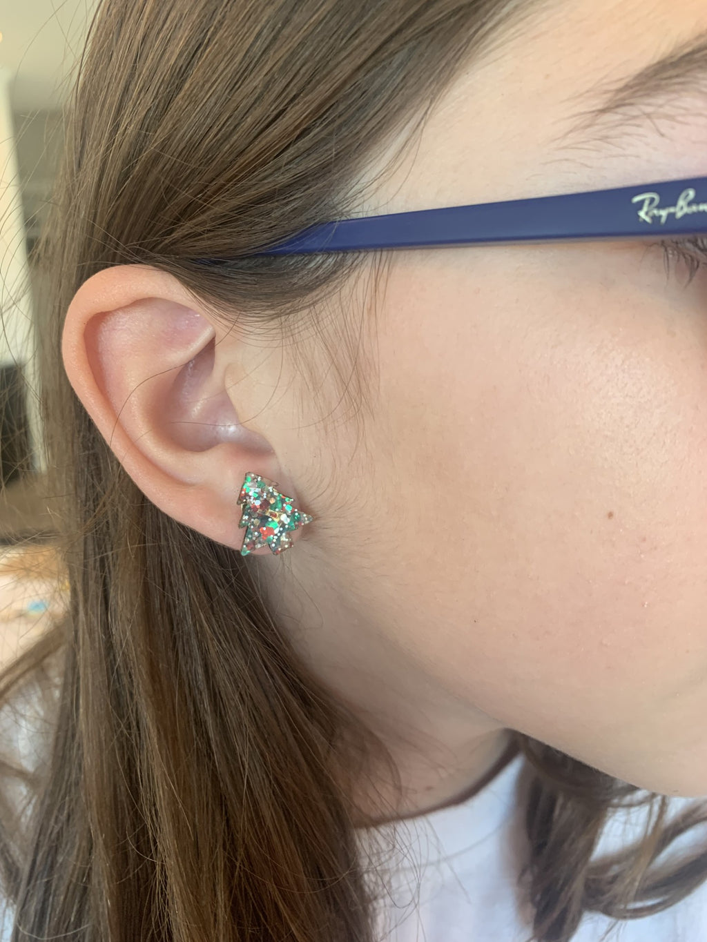 A close-up shows a person with long brown hair and blue Ray-Ban glasses wearing Christmas Tree Stud Earrings adorned with multicolored glitter. The blurred background highlights the ear and festive earrings.