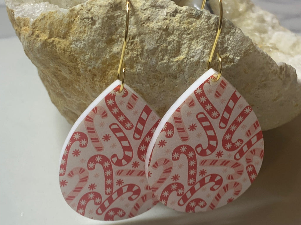 The Candy Cane Print Acrylic Earrings are teardrop-shaped with a festive red and white pattern of candy canes and snowflakes. They hang from gold hooks against a textured stone background.