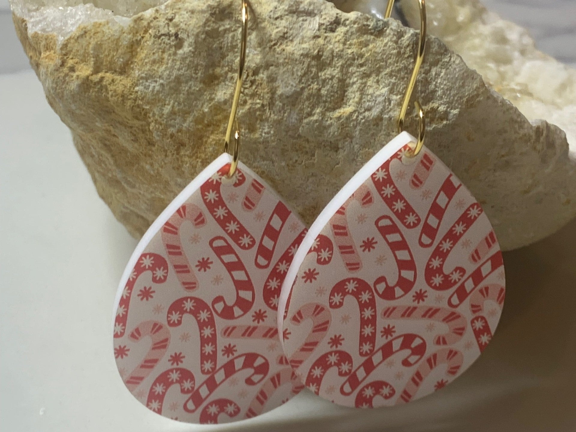 The Candy Cane Print Acrylic Earrings are teardrop-shaped with a festive red and white pattern of candy canes and snowflakes. They hang from gold hooks against a textured stone background.