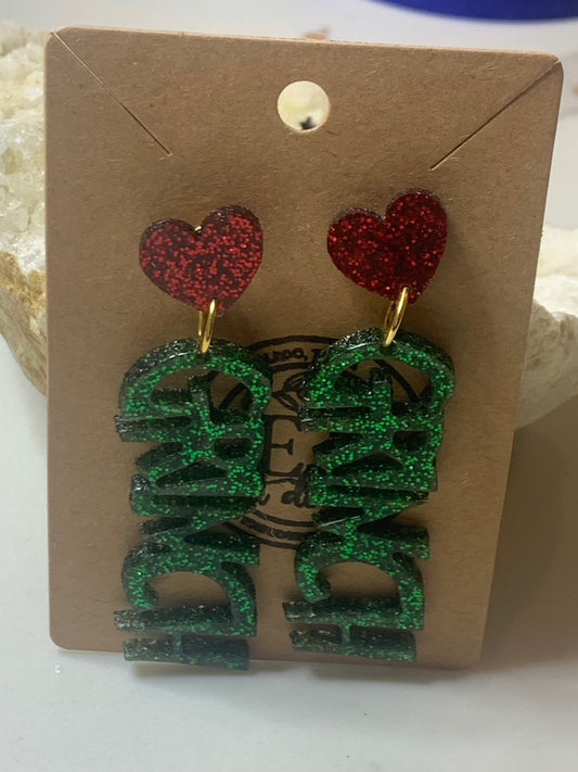 The Grinch Acrylic Earrings set includes glittery red heart studs and dangling green DAD letters, presented on a brown card. The display rests on a white surface beside a crystalline stone.