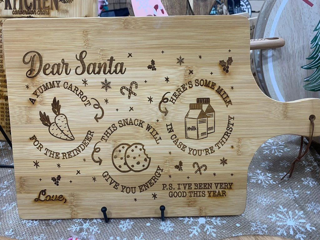 A wooden cutting board with festive engravings. Text reads: Dear Santa, a yummy carrot for the reindeer, this snack will give you energy, heres some milk in case youre thirsty. P.S. Ive been very good this year. Love, surrounded by images of a carrot, cookie, milk carton, and stars.