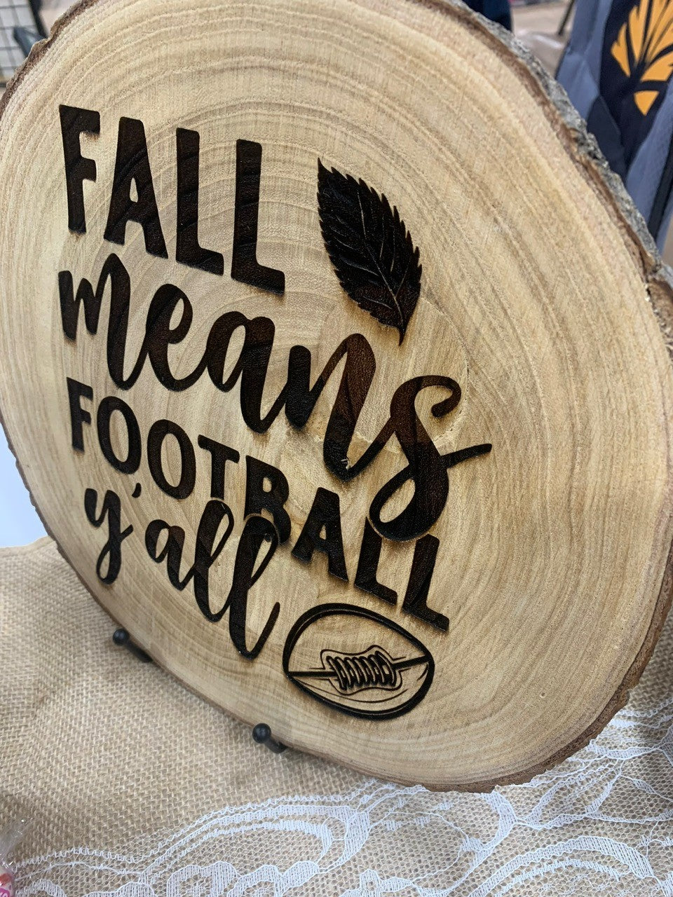 Wooden slab sign featuring the engraved phrase Fall means football yall with a leaf and a football illustration. The slab rests on a burlap surface with lace details.