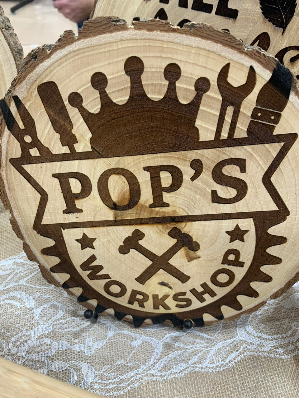 A wooden plaque, shaped like a tree slice, features a design with a large crown and various tools, such as a wrench and screwdriver. The text reads POPS WORKSHOP with two crossed hammers below. The plaque is displayed on a lace-covered table.