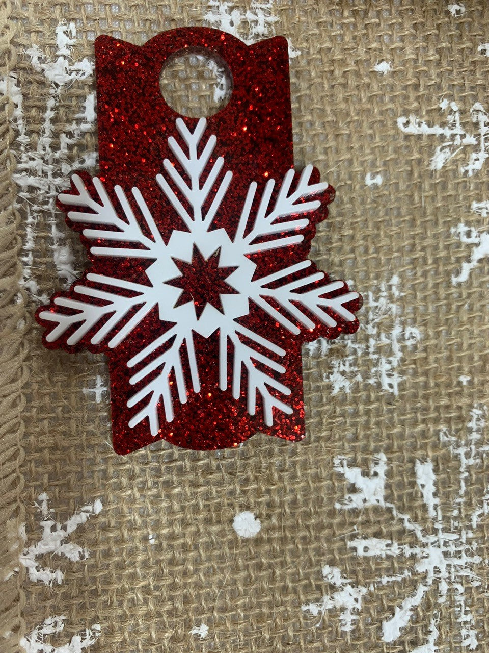 The Stanley Cup Topper Snowflake is a red glittery tag with a large white snowflake design on burlap fabric with white snowflake prints, featuring a hole at the top for hanging. This festive piece adds rustic holiday charm to any setting.