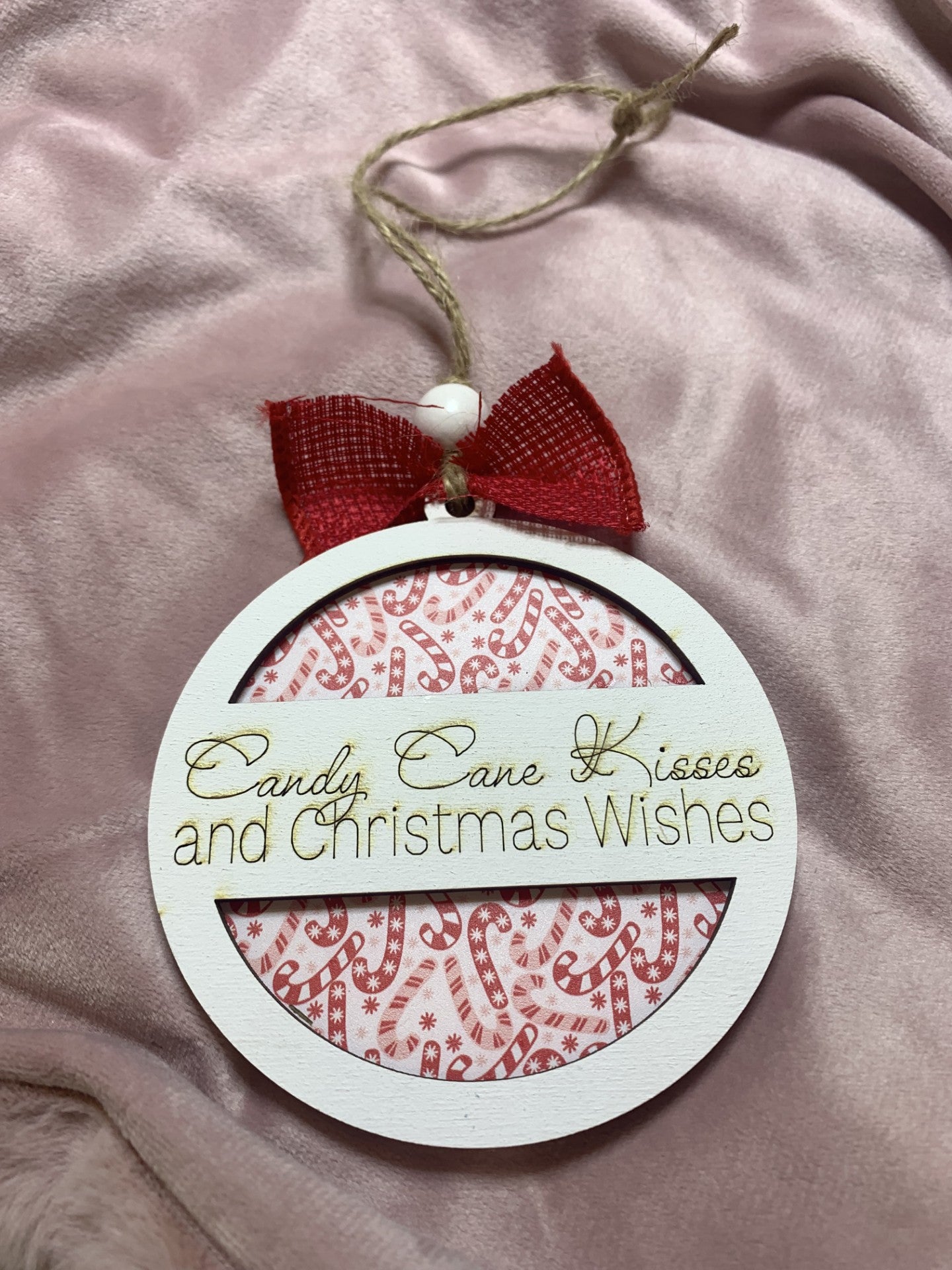The Christmas Ornaments 2 Layer Round displays a red and white candy cane pattern. Candy Cane Kisses and Christmas Wishes gleams in gold on the white border. Its adorned with a red bow and twine loop for hanging, nestled against soft pink fabric.