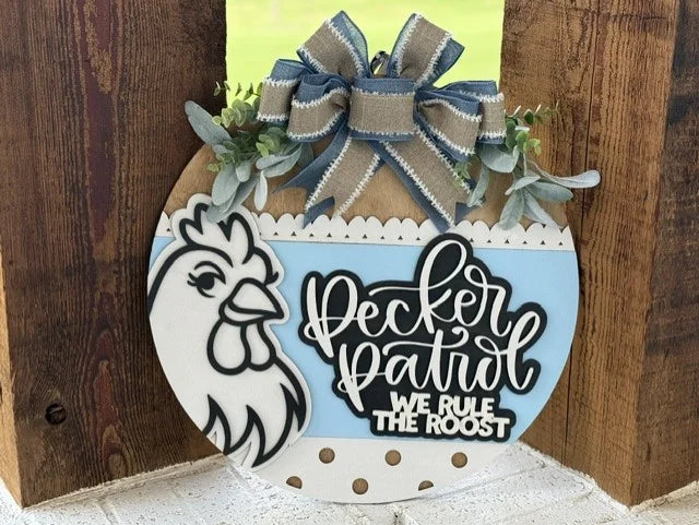 A round wooden sign with a drawing of a chicken, a large blue and burlap bow, greenery, and the text “Pecker Patrol We Rule The Roost” in white and black lettering.