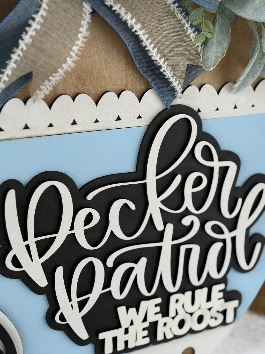 A decorative sign with layered, cursive text reading Pecker Patrol and block letters below saying We Rule the Roost, set on a light blue and scalloped background with blue and white ribbon accents.