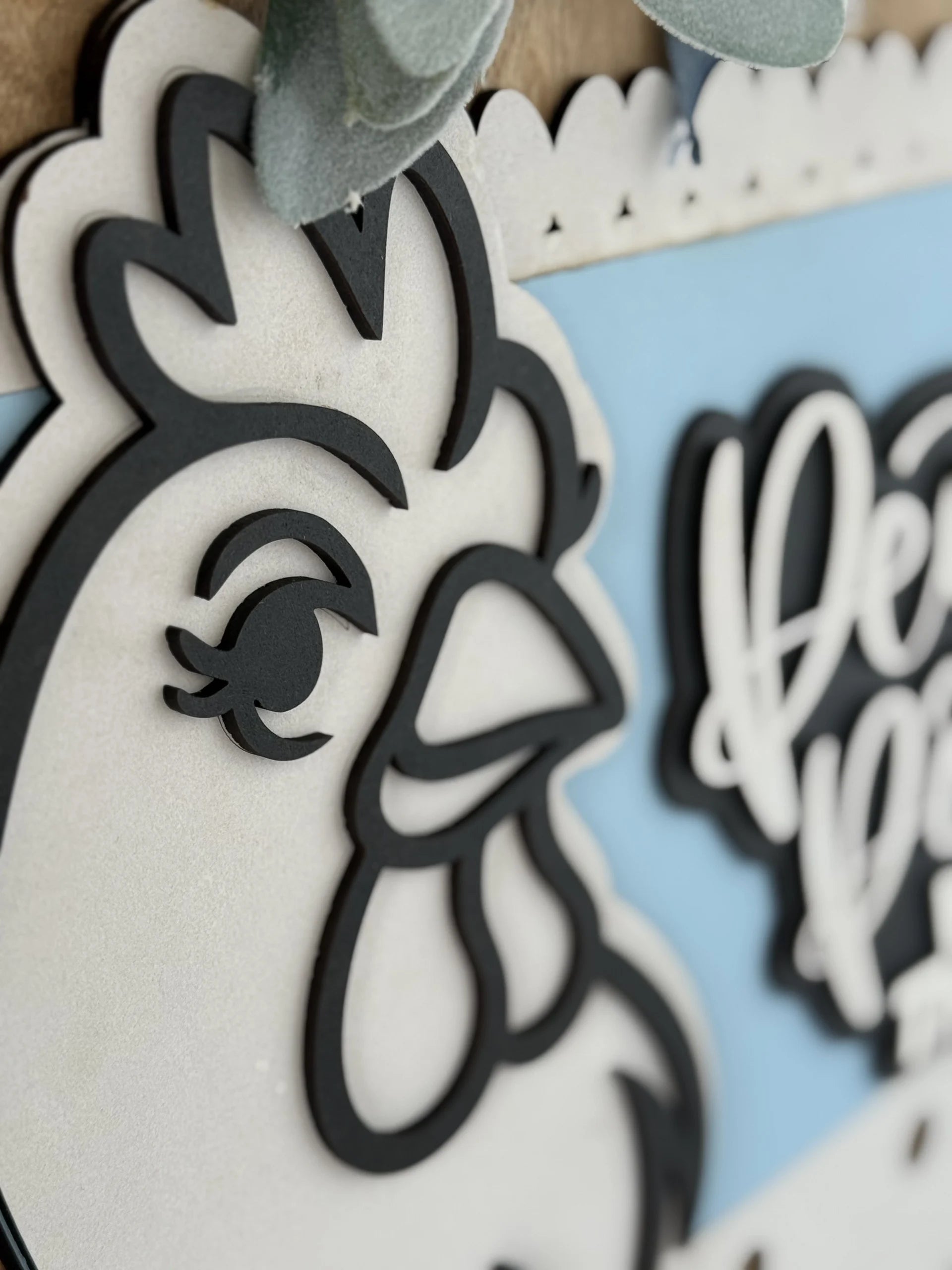 Close-up of a decorative sign featuring a stylized black and white outline of a chickens head on a blue background, with part of cursive white text visible on the right side.