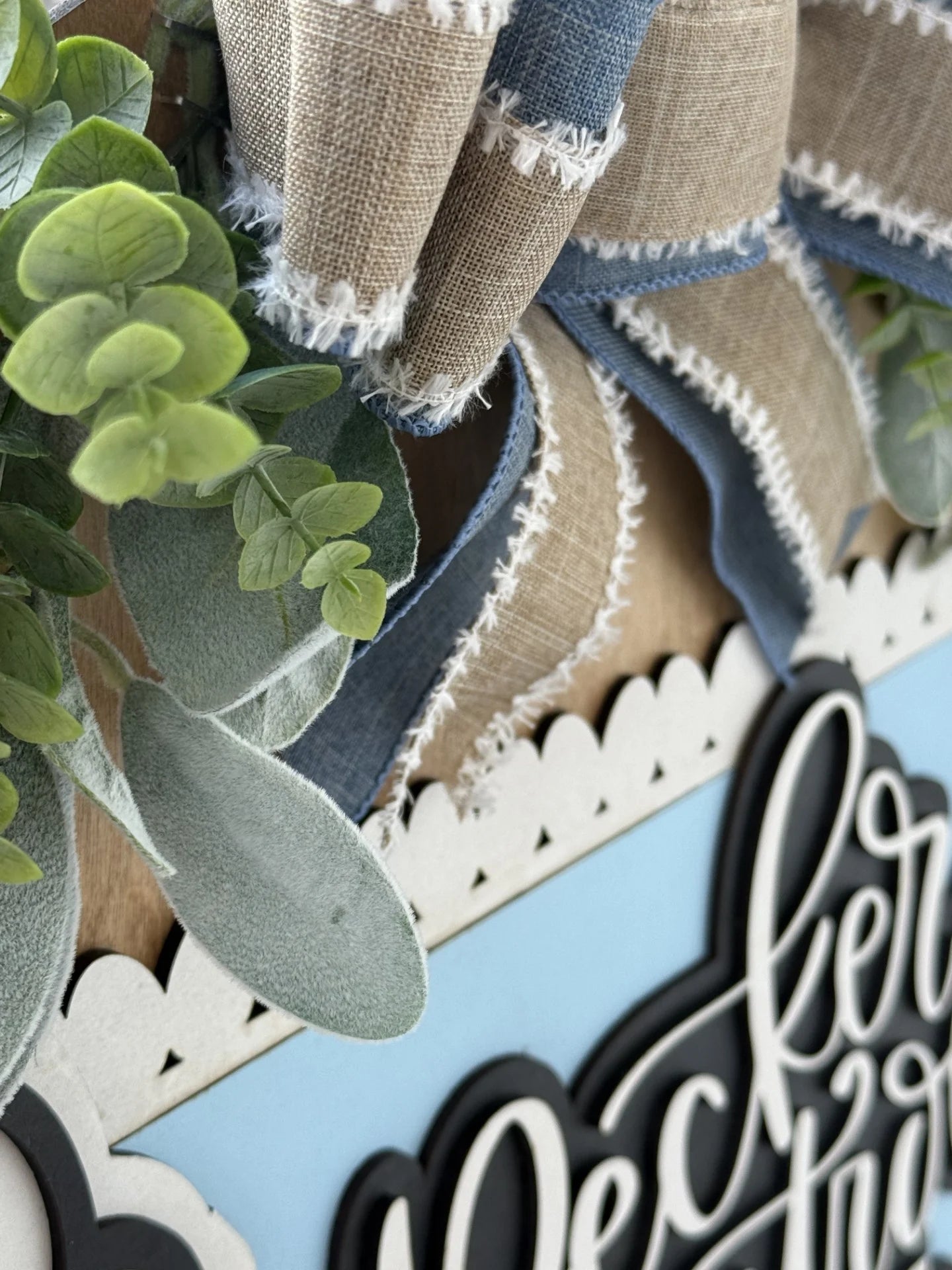 Close-up of a decorative sign featuring faux green leaves, a blue and tan burlap bow with frayed edges, and a scalloped white frame against a light blue background. Black script lettering is partially visible.