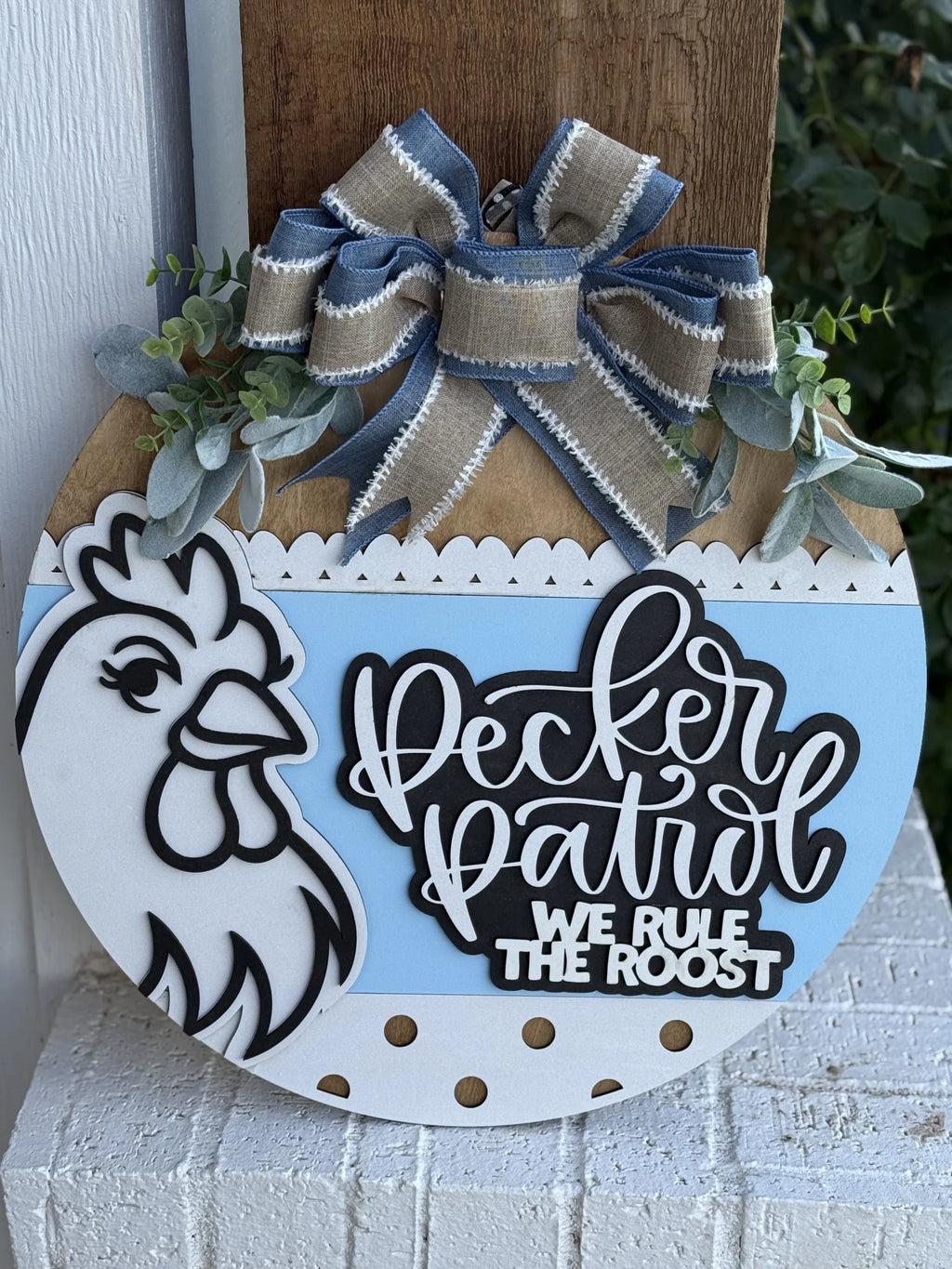 A round farmhouse sign with a black-and-white rooster, a blue and white bow, greenery, and the text Pecker Patrol WE RULE THE ROOST in bold lettering, displayed on a white brick surface.