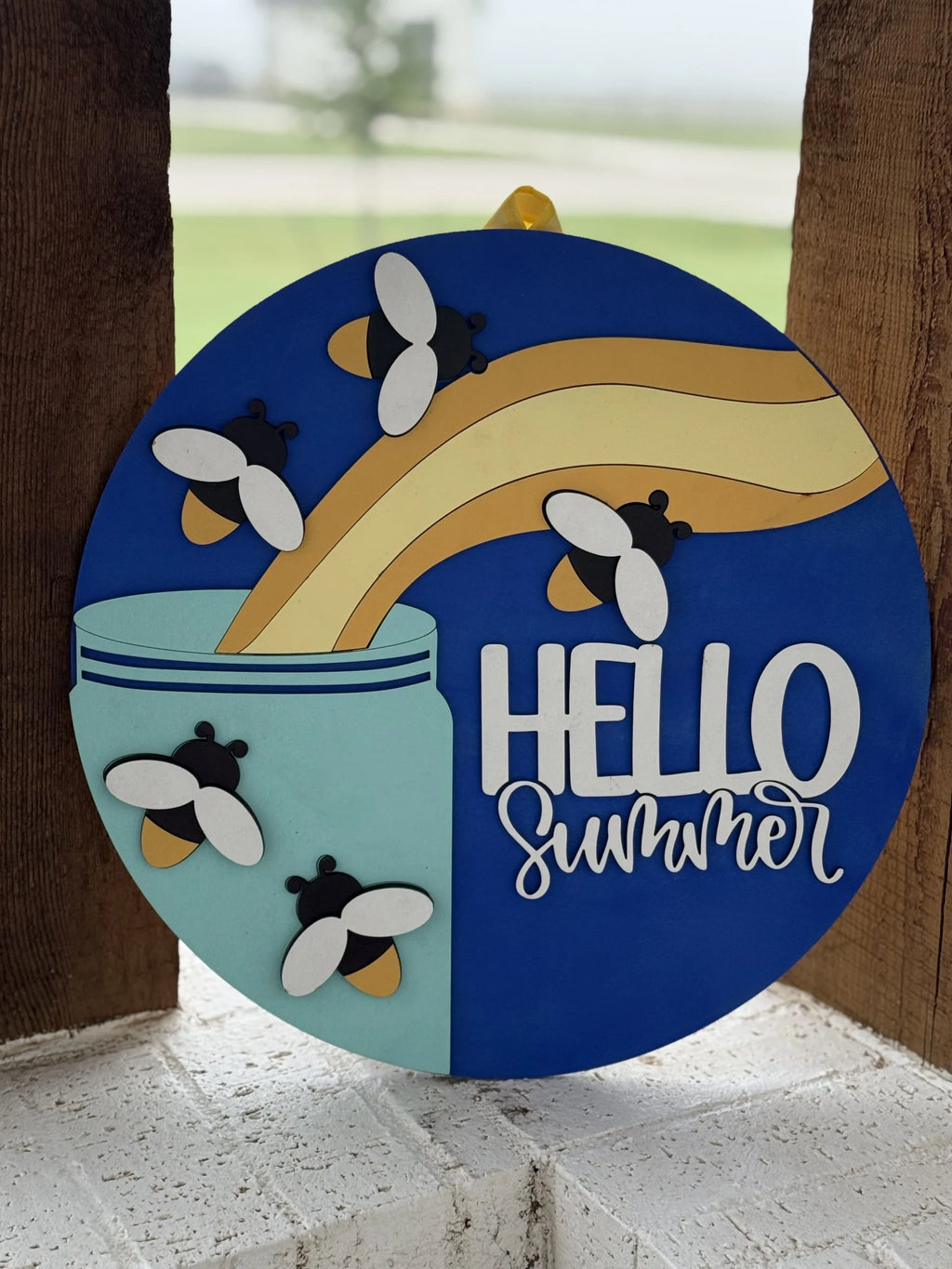 A round sign with bees flying out of a blue jar and the words HELLO Summer written in white script on a blue background. The sign is propped against a wooden surface outdoors.