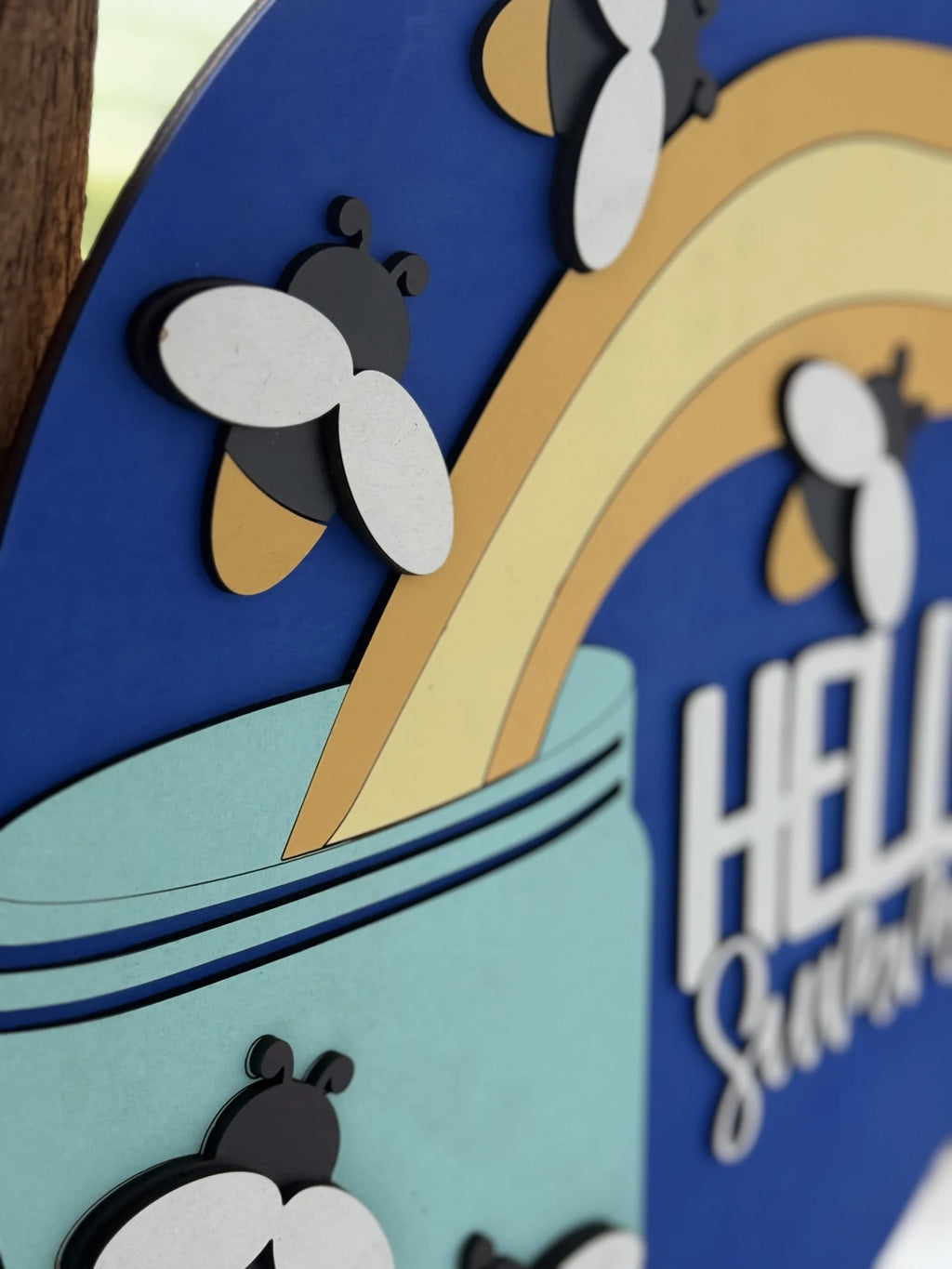 A close-up of a decorative sign with cartoon bees, a rainbow, and the partial text “HELLO” on a blue background. The design features layered, cut-out shapes for a 3D effect.