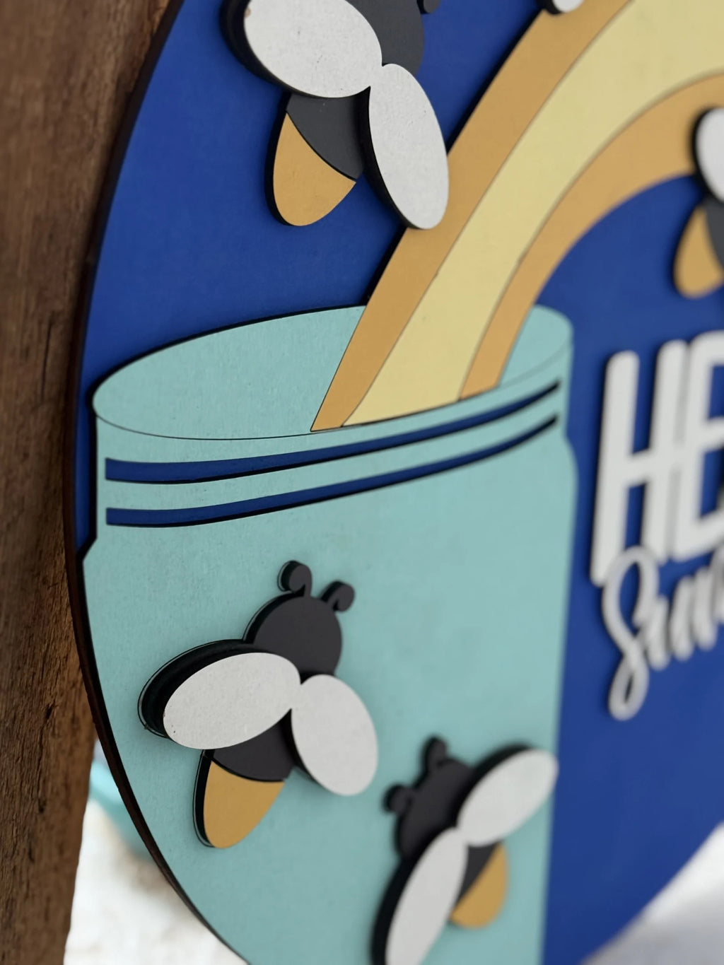 Close-up of a colorful sign featuring cartoon bees with white wings and yellow bodies, a rainbow, and a teal jar against a blue background. The layered design gives a three-dimensional effect.