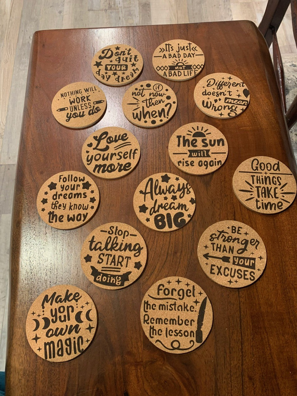 Cork coasters with motivational quotes are laid out on a wooden table. Phrases include: Don’t quit your day dream, The sun will rise again, Always dream big, and Forget the mistake, remember the lesson, each in decorative fonts and surrounded by small stars.