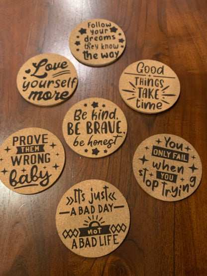 A set of four motivational cork coasters is displayed on a wooden surface, featuring phrases like Follow your dreams and Good things take time. These coasters inspire with every sip, reminding you that You only fail when you stop trying.
