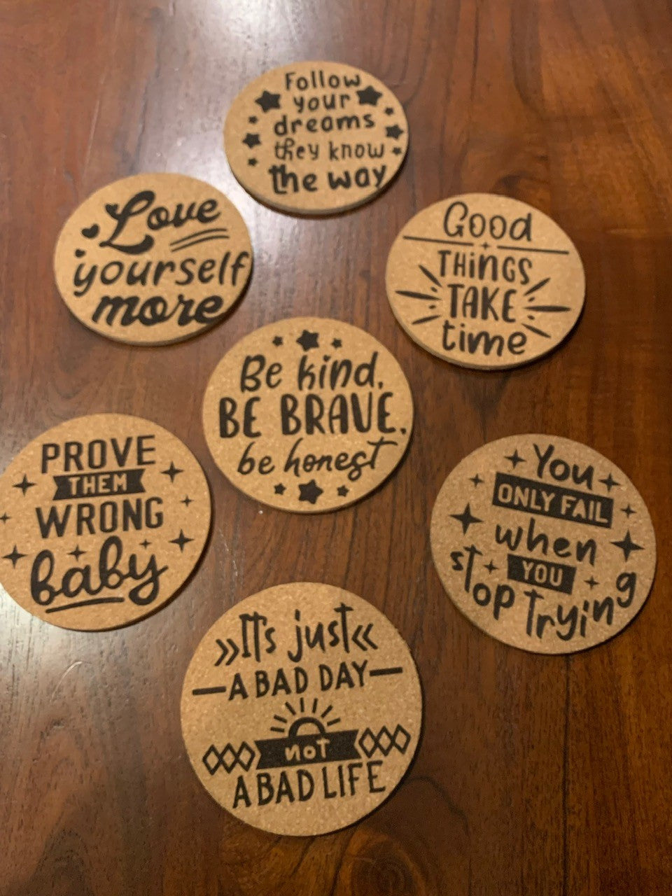 Seven round coasters with motivational quotes are arranged on a wooden surface. Phrases include Love yourself more, Follow your dreams, they know the way, and Good things take time. Each coaster features unique typography and decorative elements like hearts and stars.