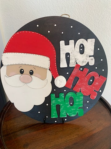 The Ho Ho Ho Santa Door Sign features a cheerful cartoon Santa in a red hat with white trim, alongside colorful Ho Ho Ho text on a snowy black background. This delightful piece is perfect for enhancing your holiday decor and hangs elegantly on any door.