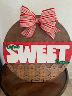 The Home Sweet Home Gingerbread Door Hanger displays a red and white striped bow on a round wooden sign. SWEET stands out in white with smaller green home letters. Gingerbread figures line the bottom against alternating red and natural wood, making it ideal for festive decor!.