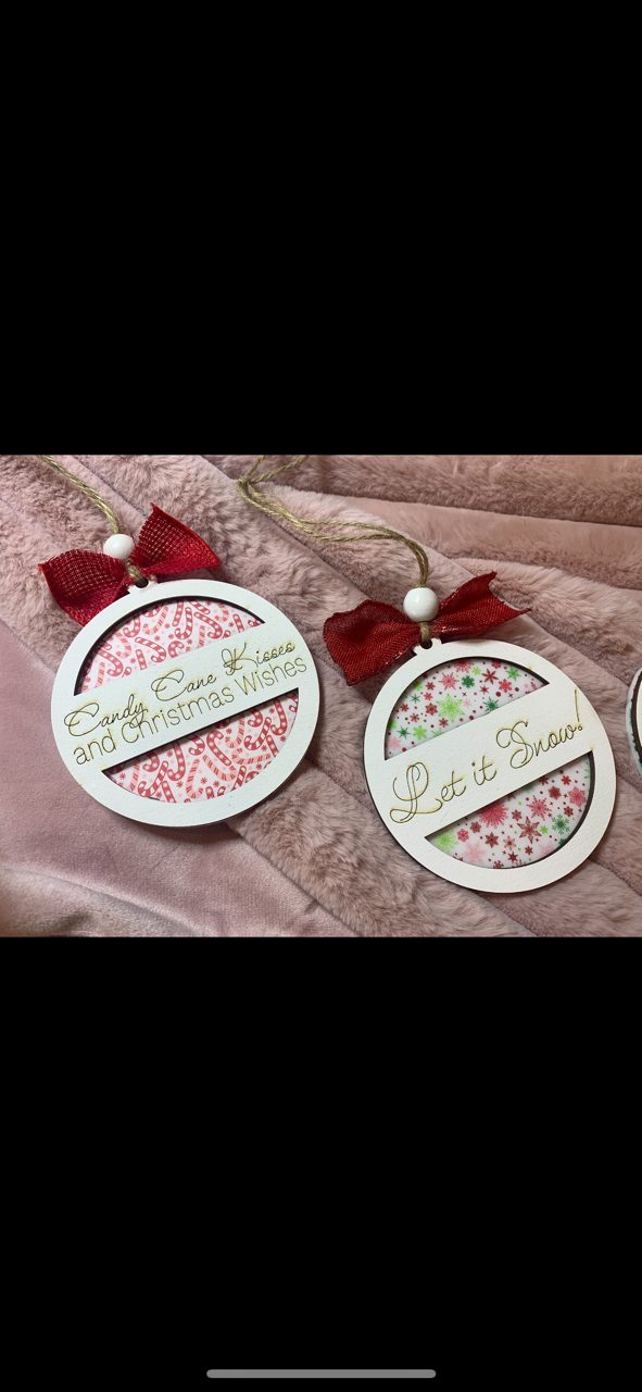 Two round Christmas ornaments with red bows. The left ornament says Candy Cane Wishes and Christmas Kisses in cursive with a red and white pattern inside. The right ornament says Let it Snow! with a green and red snowflake design inside, lying on a soft pink blanket.