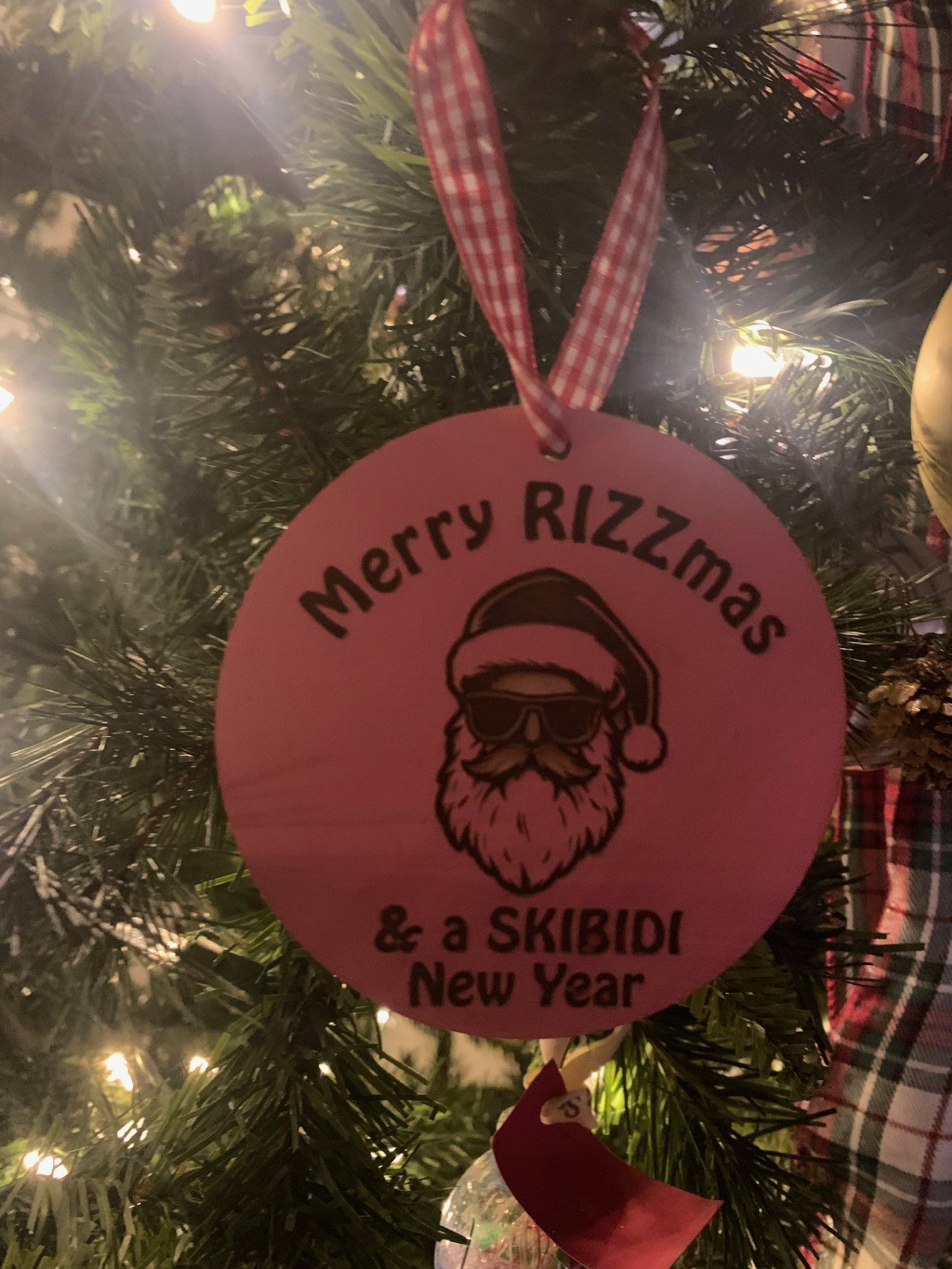 The Merry RIZZmas & a SKIBIDI New Year Santa Christmas Ornament - Pink, featuring a bearded man in a Santa hat and sunglasses, hangs on the tree amid twinkling lights and lush pine needles.