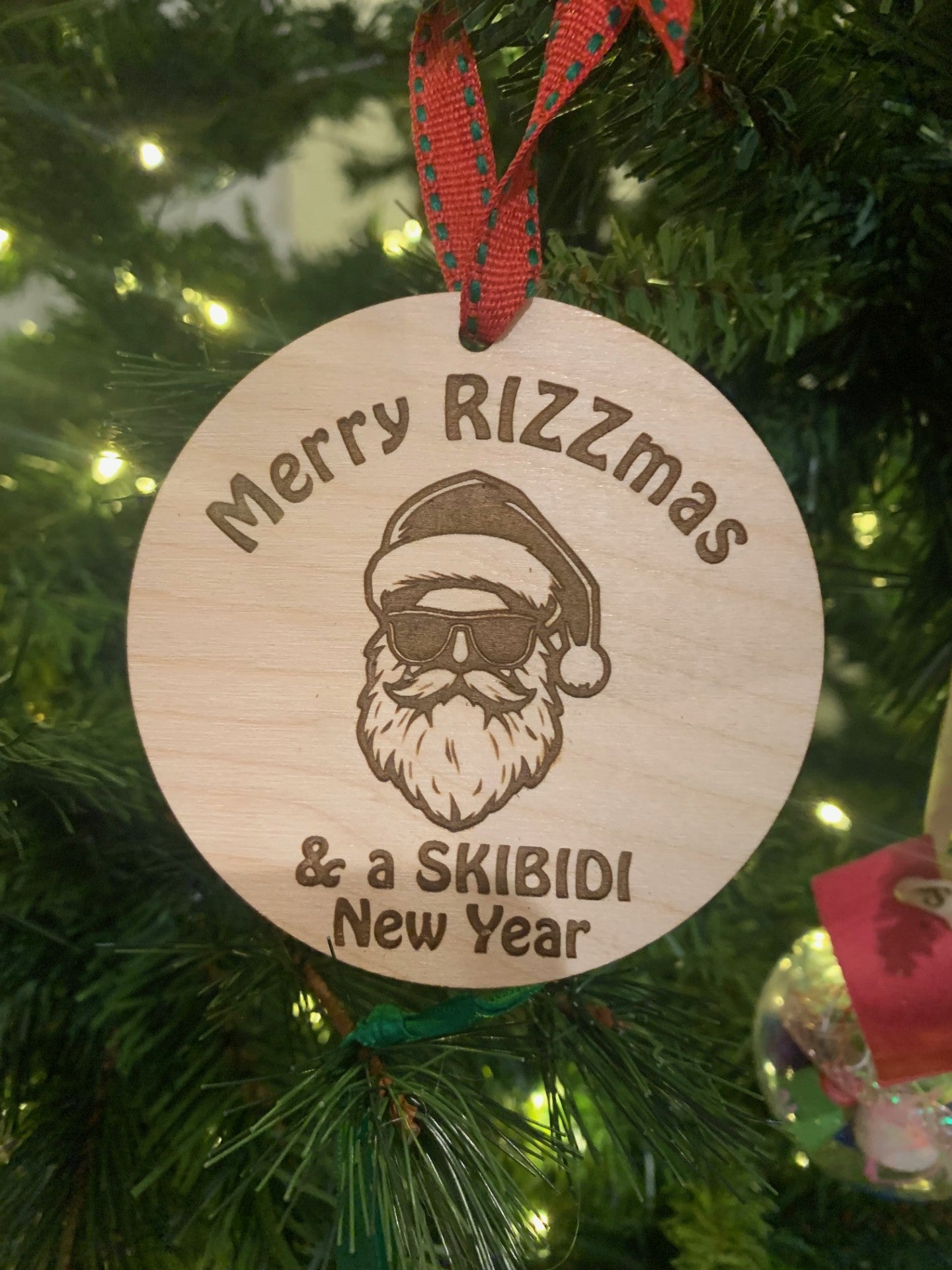 The Merry RIZZmas & a SKIBIDI New Year Santa Christmas Ornament features a bearded Santa in sunglasses and hat, engraved with festive wishes. It hangs by a red ribbon on the tree, surrounded by twinkling lights.