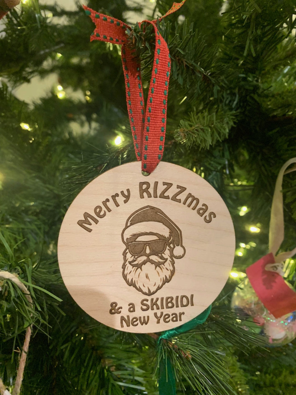 The Merry RIZZmas & a SKIBIDI New Year Santa Christmas Ornament features Santa with sunglasses and a hat, etched with festive text. Its adorned with a red and green ribbon, making it a charming addition to your holiday decor.