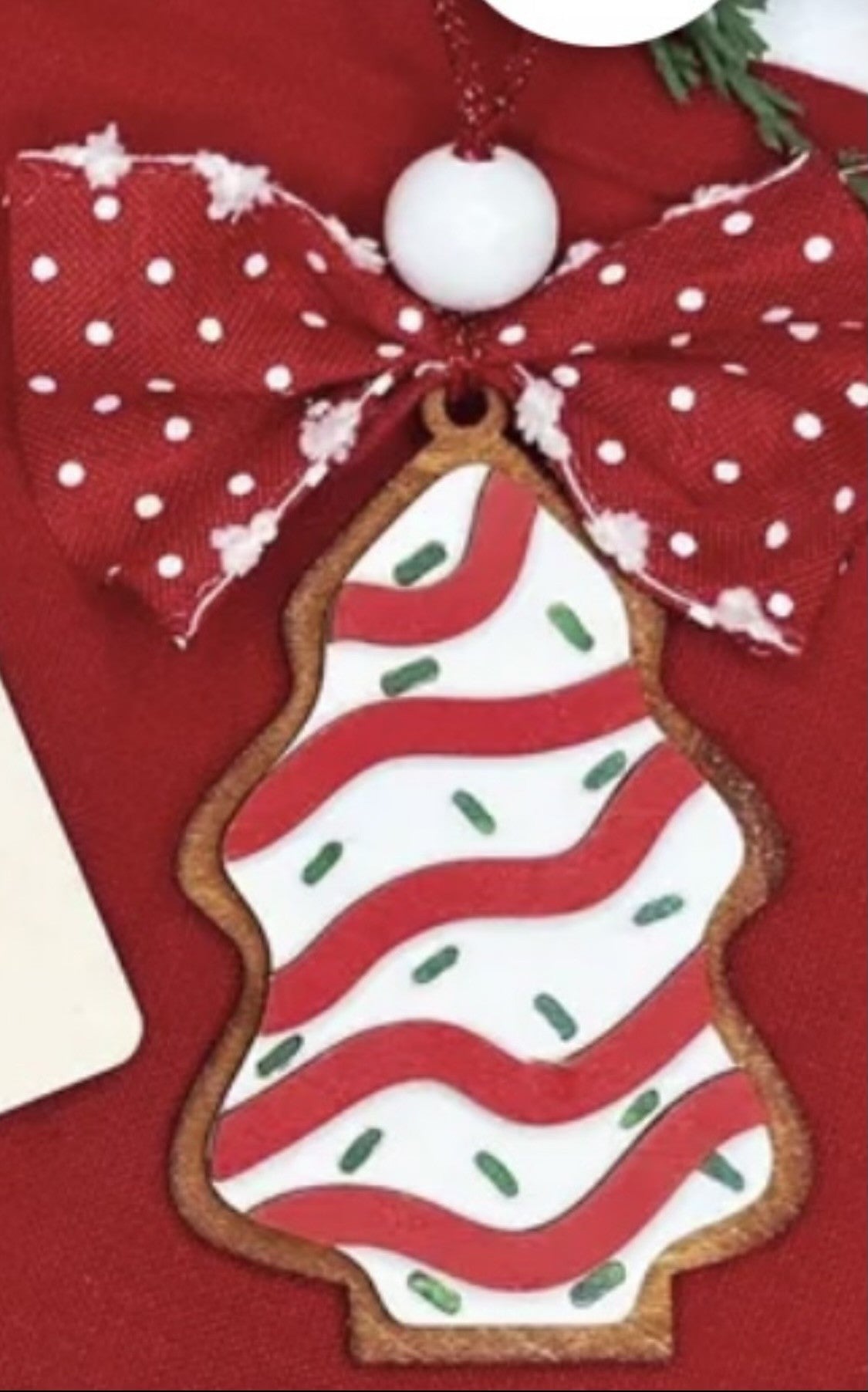 A Christmas tree-shaped cookie ornament with red and white stripes and green sprinkle-like decorations. It is topped with a red and white polka dot bow and hangs by a string. The background features a red surface, enhancing the festive theme.