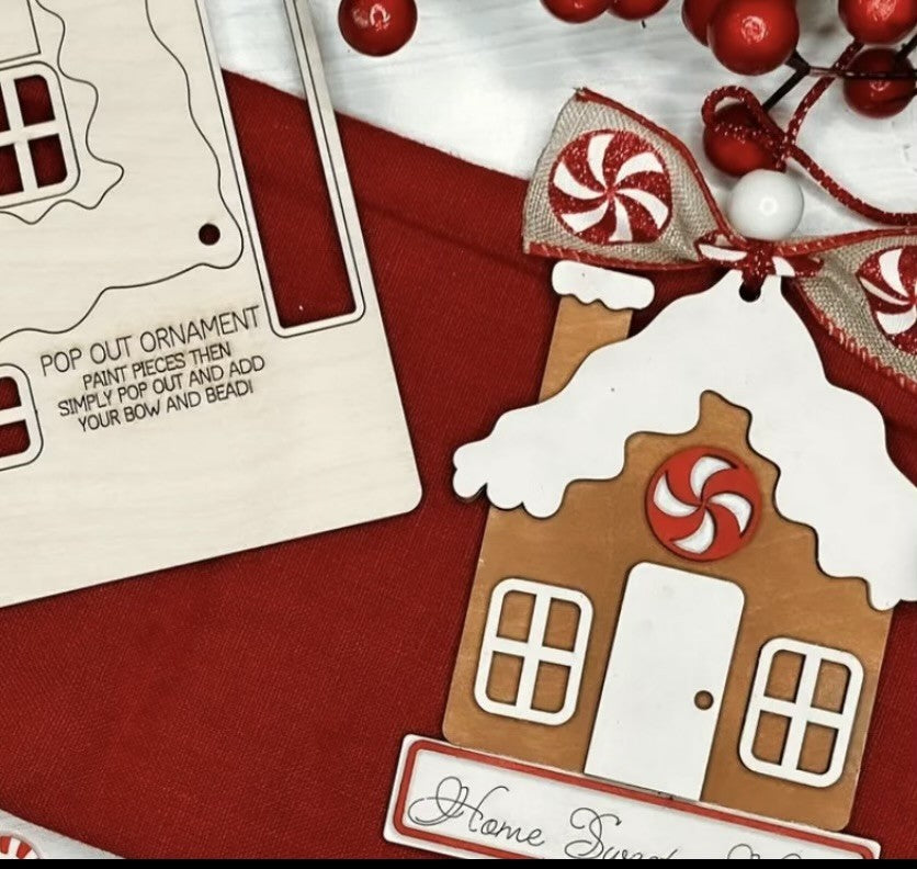 A wooden gingerbread house ornament is on a red cloth. It features a peppermint swirl above the door, with instructions nearby on how to assemble. A cluster of red and white berries with ribbon is in the top corner, contributing to the festive theme.