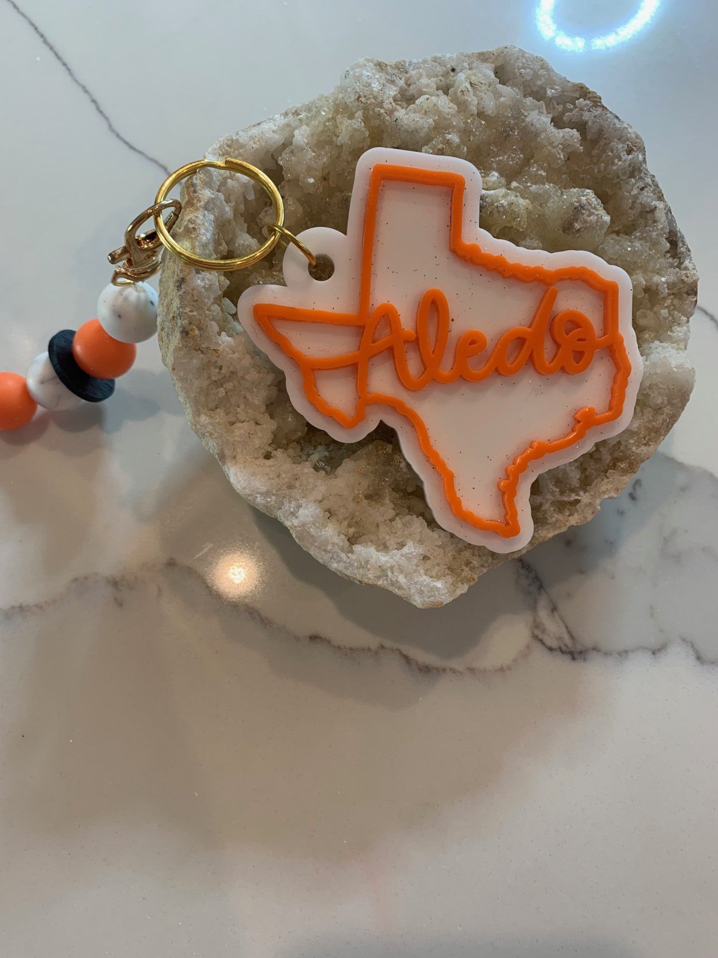 The Aledo Texas Keychain with Beads, shaped like Texas and displaying Aledo in vibrant orange, rests on a rock with black, orange, and white beads. It sits against a sleek marble background, capturing the charm of Aledo.