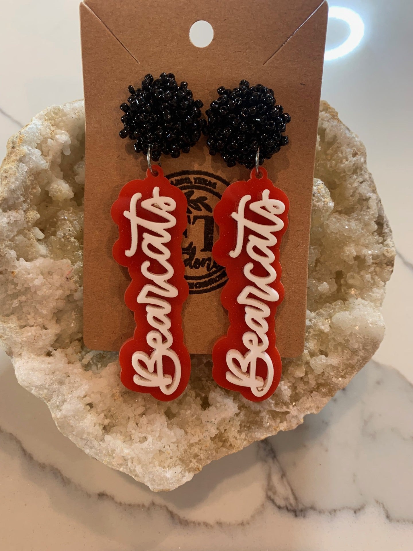 The Aledo Bearcat Earrings feature black beaded studs with red and white acrylic pendants spelling Bearcats, displayed on a brown card with a branded logo, elegantly set against a textured stone backdrop.
