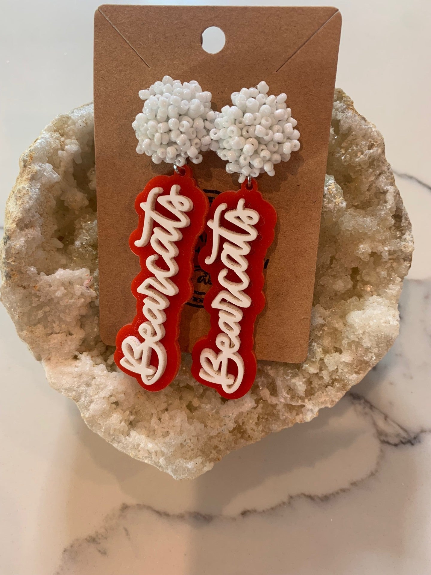 Displayed on a textured beige stone background, the striking Aledo Bearcat Earrings feature a white beaded top and red-and-white dangles with Bearcats. Elegantly attached to a brown card, they are a perfect homage to Aledo spirit.