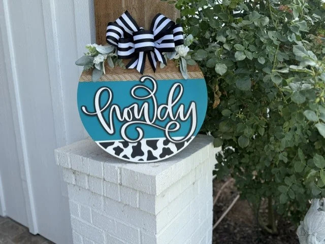 A round sign decorated with a black and white striped bow and greenery reads howdy in script on a teal and cow-print background, resting on a white brick column near green bushes.