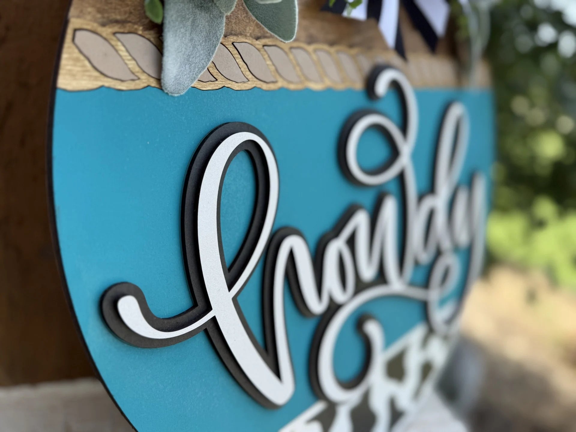 Close-up of a decorative sign with raised, cursive text that reads Howdy on a teal and wood background. The top of the sign is adorned with artificial greenery and a black-and-white ribbon.