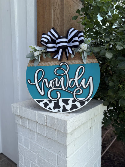 A round sign with the word howdy in script, decorated with a black and white striped bow, greenery, and a cow-print pattern, hangs on a white brick post near a leafy plant.