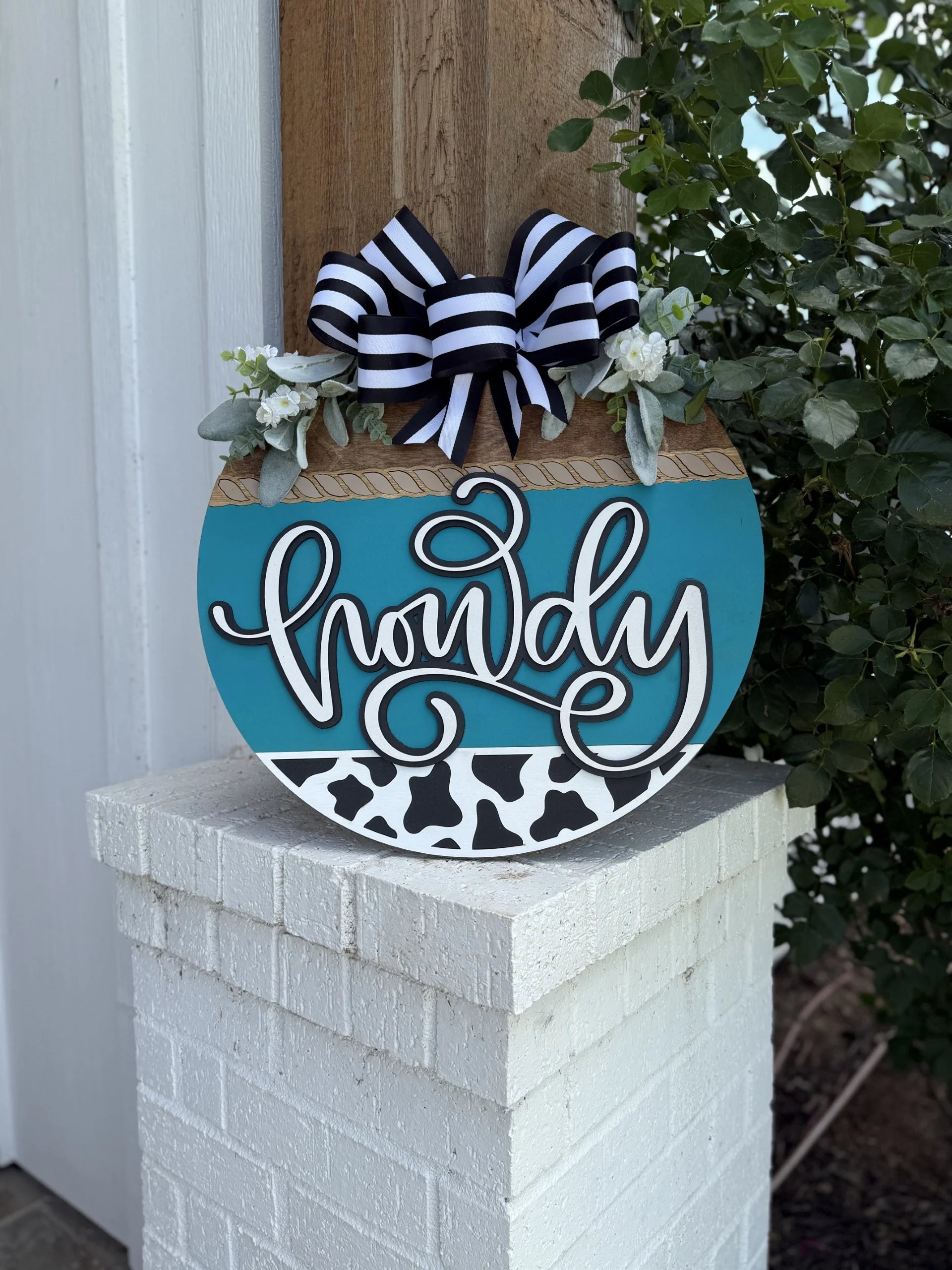A round sign with the word howdy in script, decorated with a black and white striped bow, greenery, and a cow-print pattern, hangs on a white brick post near a leafy plant.