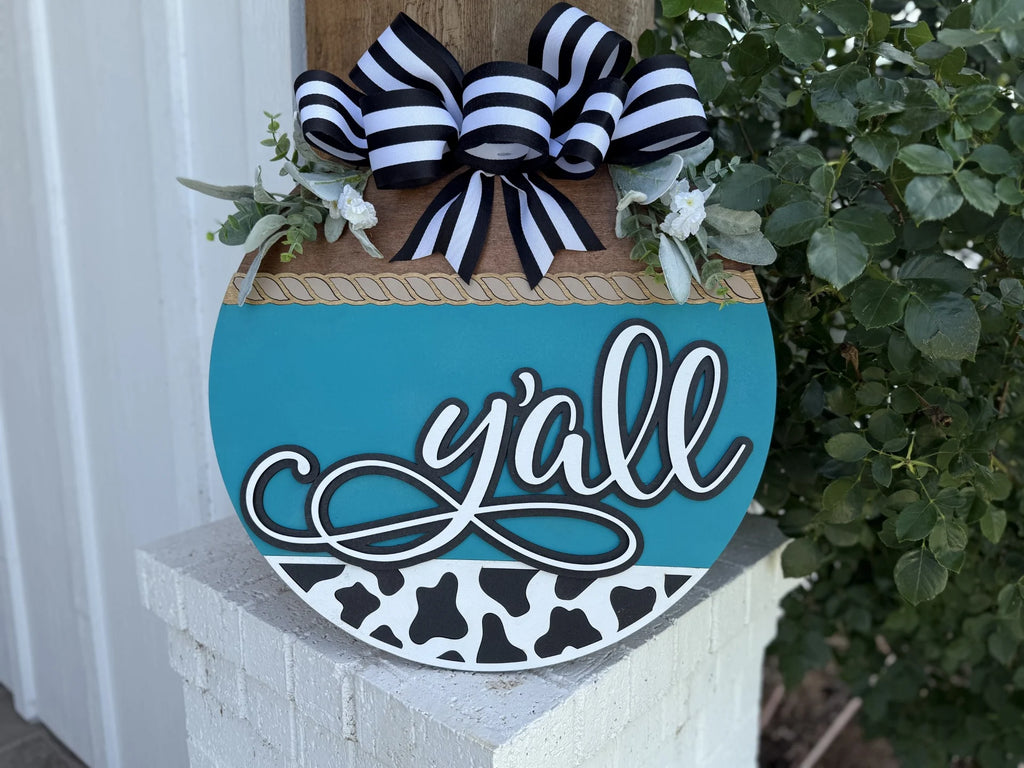 A round decorative sign with “y’all” written in script, featuring a teal and cow-print design, topped with a black-and-white striped bow and greenery, hanging on a white porch post next to green leaves.