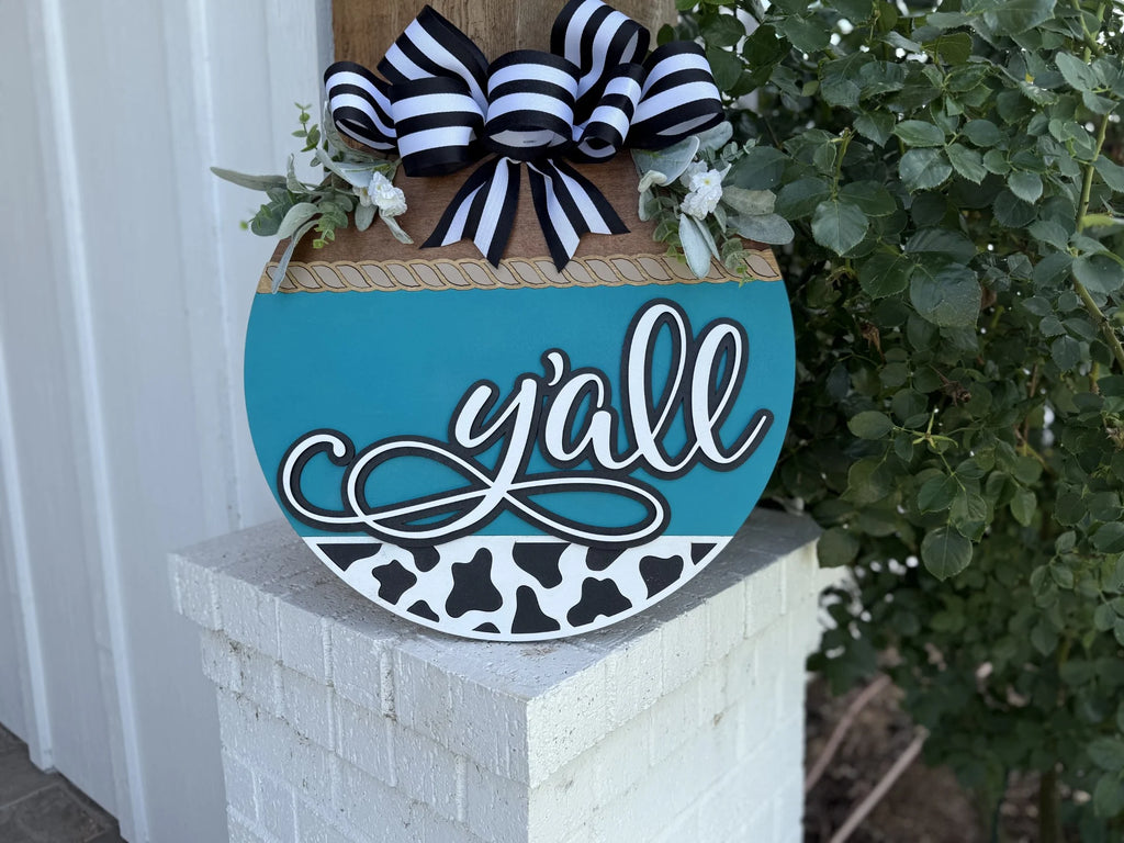 A round blue and white sign with yall written in script, decorated with black and white striped bows, greenery, and a cow print pattern at the bottom, sits on a white brick pillar near leafy plants.